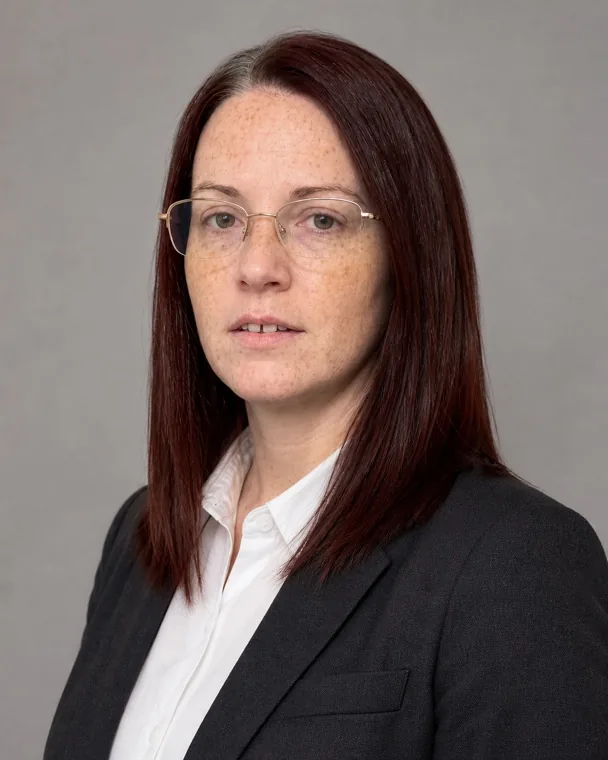 Nadia in a corporate headshot: light grey studio background, charcoal suit over a white shirt, the same freckles, grey streak and wire glasses