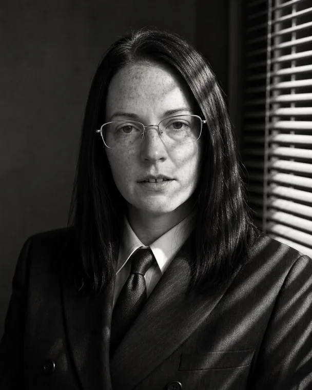 Nadia in a black-and-white film noir portrait, hard light through window blinds, the same glasses and freckles