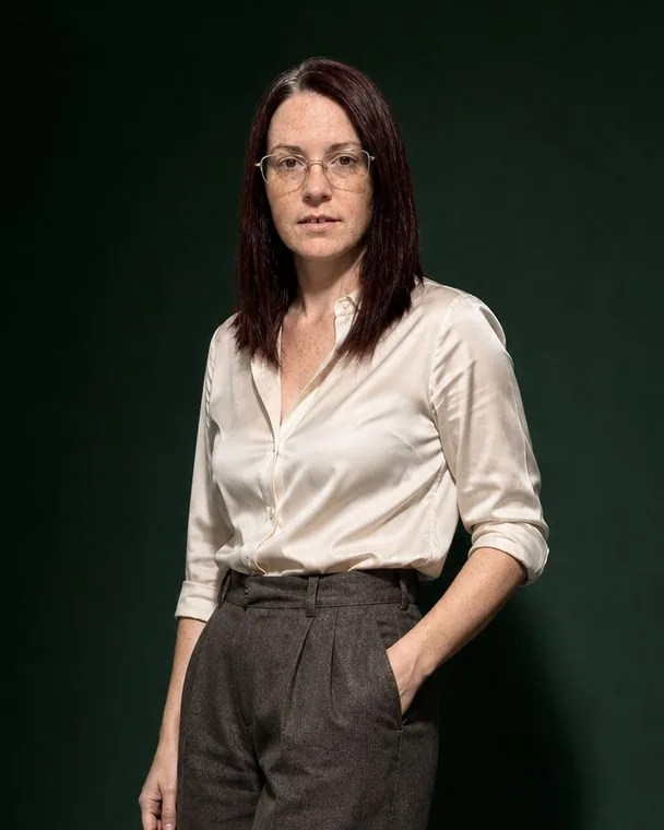 Nadia standing against a deep green studio backdrop, cream silk shirt tucked into wide brown wool trousers, one hand in a pocket, the same glasses and freckles