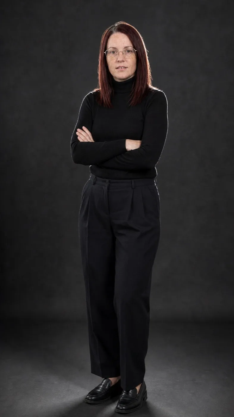 Nadia head to toe in a black roll-neck and wide trousers against a dark studio wall, arms folded, same glasses and grey streak