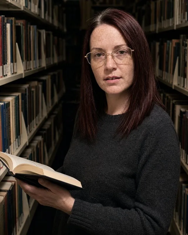 Author: standing among library shelves with a book half open, charcoal wool jumper, deep shadows between the stacks