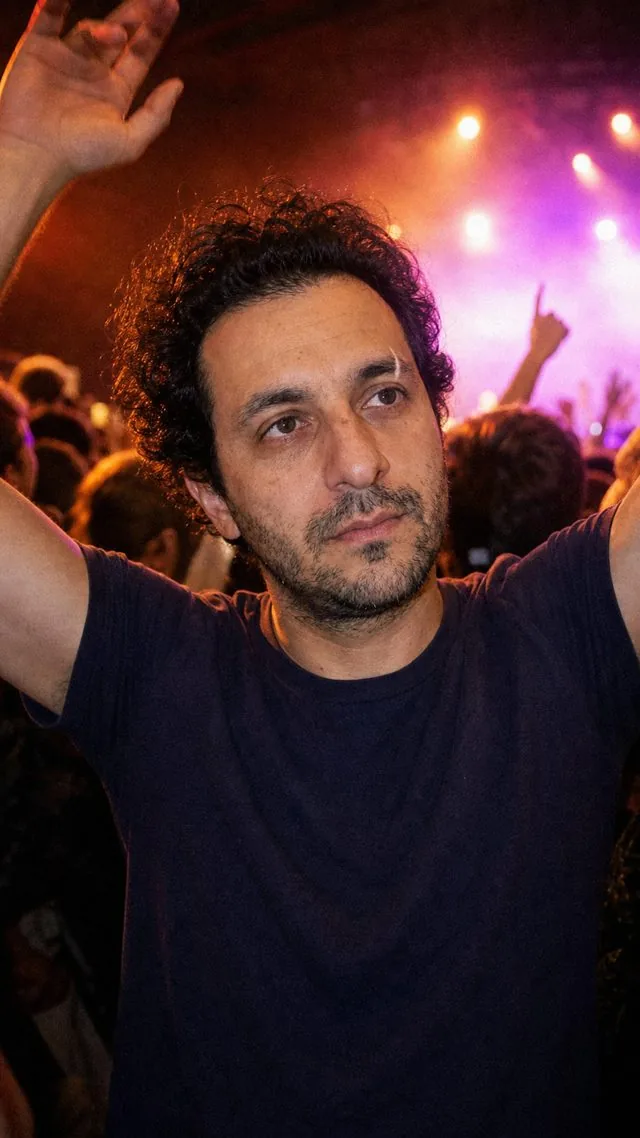 Concert: in a concert crowd with arms half raised, stage lights flaring behind, deep colour and heavy grain
