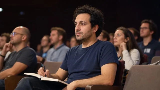 Conference: seated in an audience with a notebook, stage light spilling from the front of the room