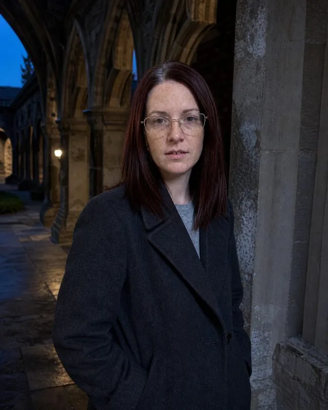 Dark academia: in a stone cloister at dusk in a long coat, cold blue light and long arches behind