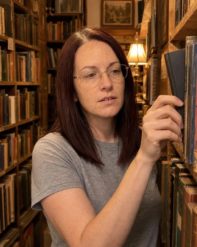 Bookshop: browsing a shelf in an old bookshop, one book half pulled out, warm lamp light between narrow stacks