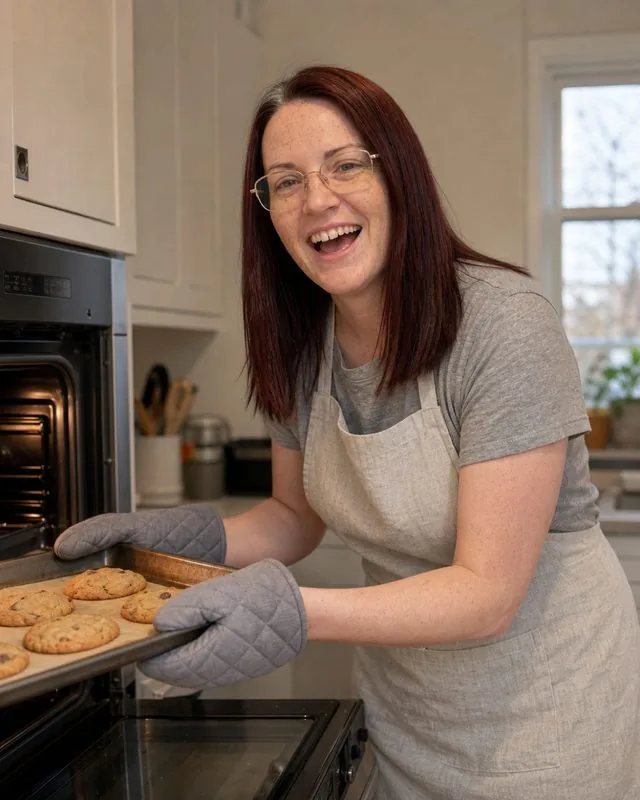 Cooking at home: pulling a tray from the oven in an apron, mid-laugh, soft window light from the left