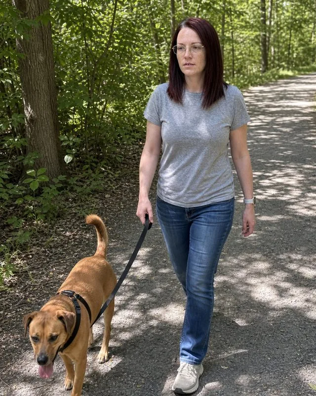With a dog: walking a dog on a lead along a tree-lined path, candid mid-stride, dappled light through leaves
