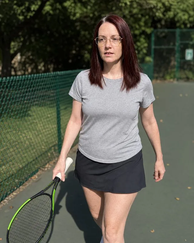 Tennis club: walking off court with the racket swinging, green fencing and trees out of focus behind