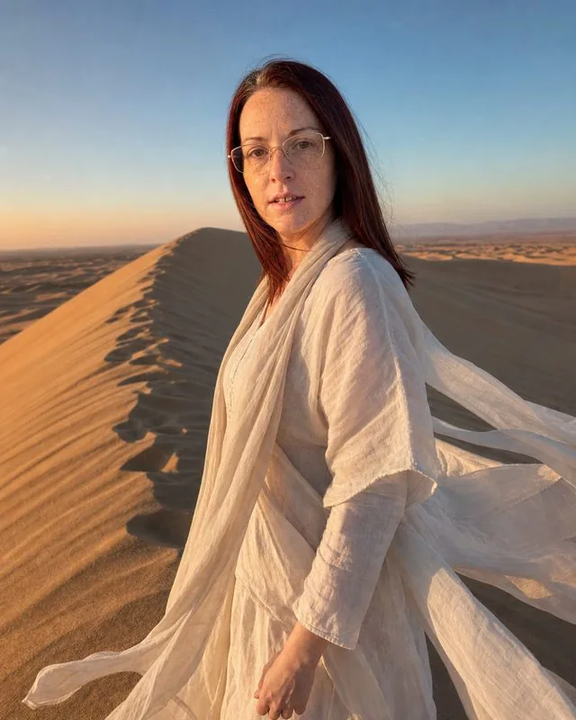 Desert dunes: on a high sand ridge in flowing light clothing, wind lifting fabric, hard low sun and long shadows