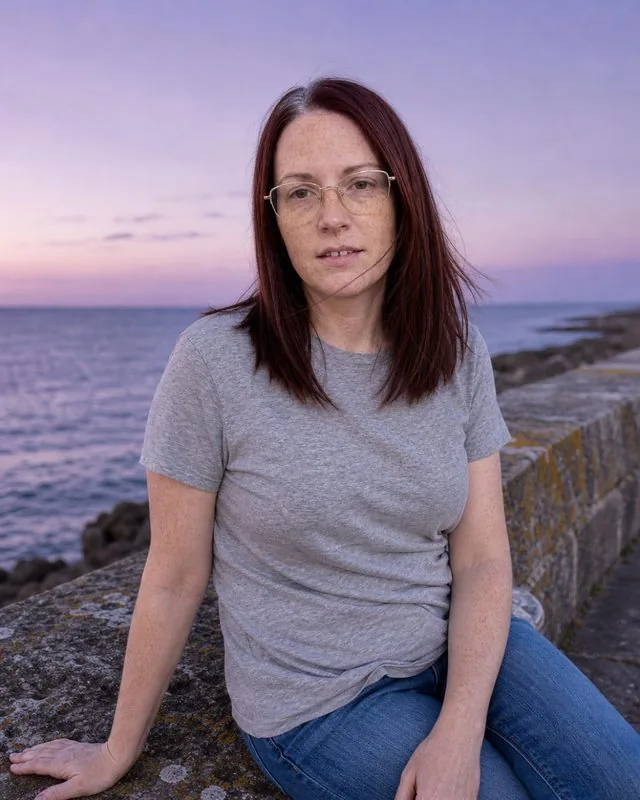 Engagement shoot: seated on a stone wall by the sea at dusk, wind in the clothes, soft violet sky