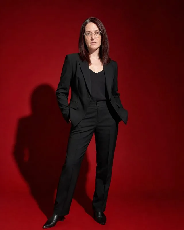 Editorial: a full-length frame against a deep red backdrop, sharply cut black suit, strong pose