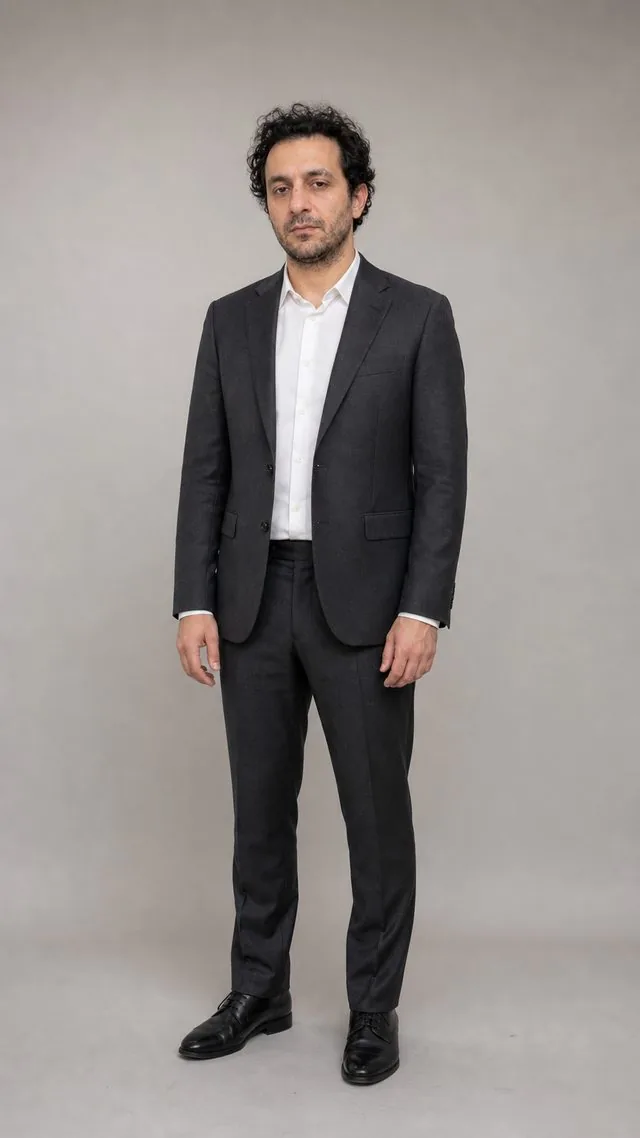 Business full-length: a full-length frame from head to toe against a light grey studio wall, charcoal wool suit over a white shirt