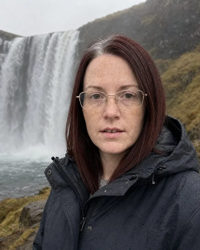 Iceland: beside a wide waterfall with spray in the air, heavy waterproof jacket with the hood down, cool flat light