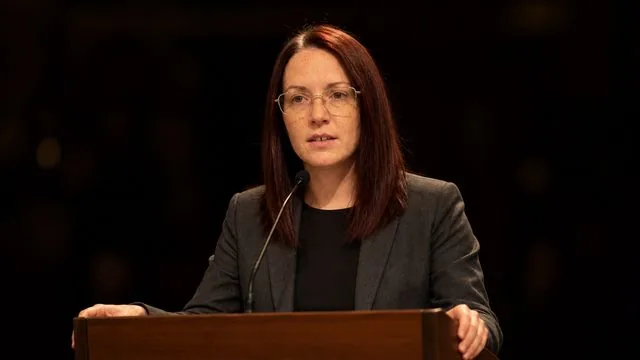 Keynote speaker: standing at a lectern under warm stage wash, hands on the edges of the lectern, dark auditorium behind