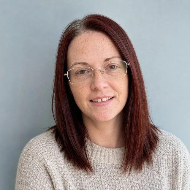 LinkedIn: a square crop against a pale blue wall, light knit jumper, open smile, soft window light from the right
