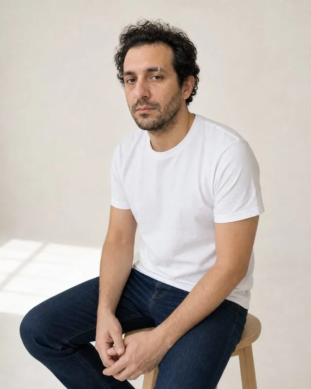 Minimal studio: seated on a pale wooden stool in an empty studio, plain white shirt, large diffused window light from one side