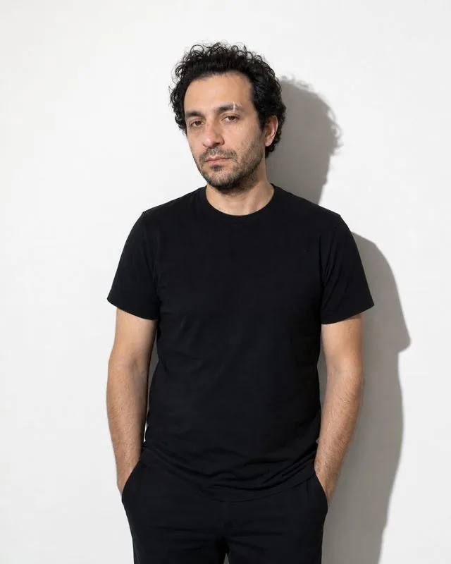 Minimal studio: a standing frame in simple black clothing, single hard light casting a crisp shadow on a white wall