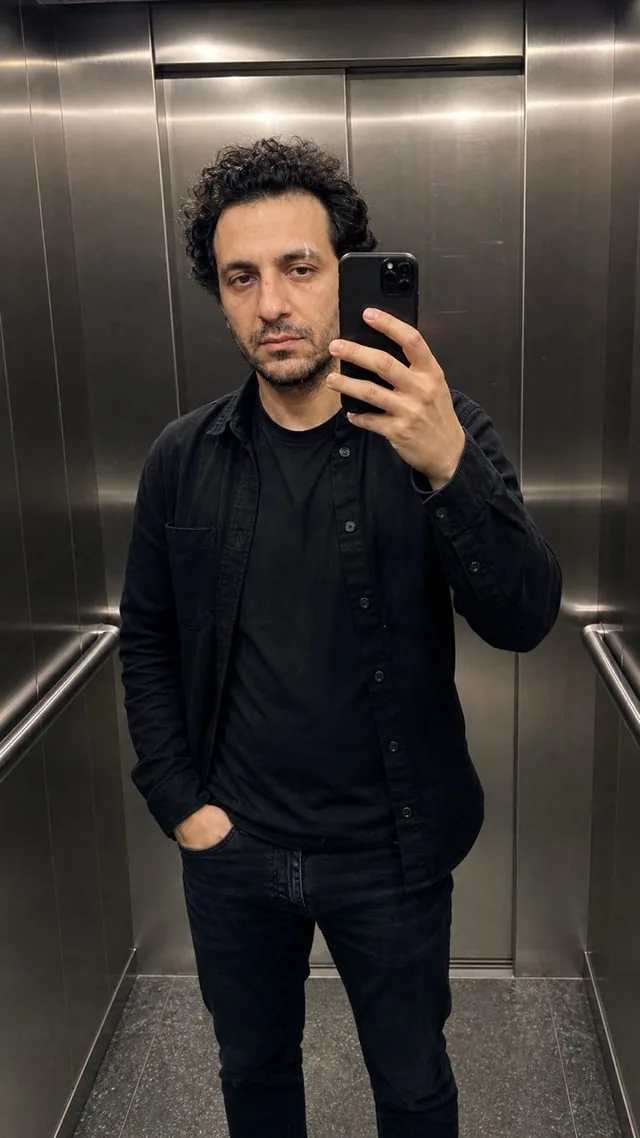 Mirror selfie: a mirror selfie in a lift with brushed metal walls, black overshirt and dark jeans, overhead light