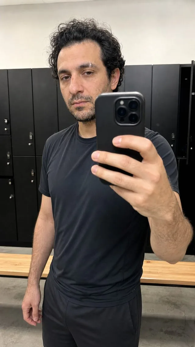 Mirror selfie: a gym changing-room mirror selfie in athletic clothing, cool fluorescent light, phone half covering the frame