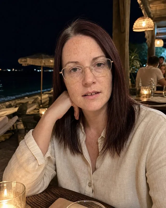 Mykonos nights: at a beach club table with candles and low warm lamps, cream linen shirt, sea dark behind