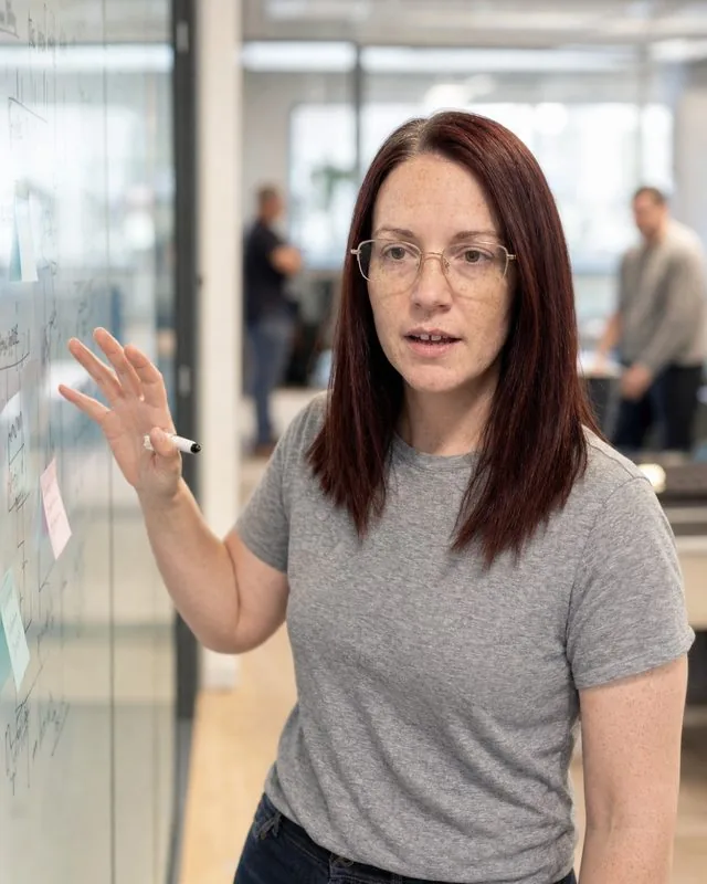 Office candid: standing at a glass meeting-room wall covered in notes, mid-gesture, colleagues blurred well behind