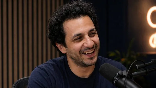 Podcast host: mid-laugh in a podcast studio with acoustic panels and a neon sign glowing softly out of focus behind