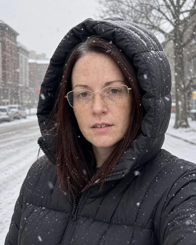 Puffer jacket: in an oversized puffer jacket on a snowy street, hood up, cold flat daylight and falling flakes