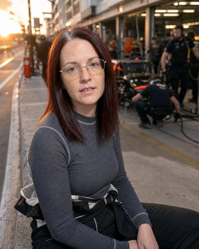 Race driver: seated on a pit wall with the team behind, racing suit with the sleeves tied at the waist