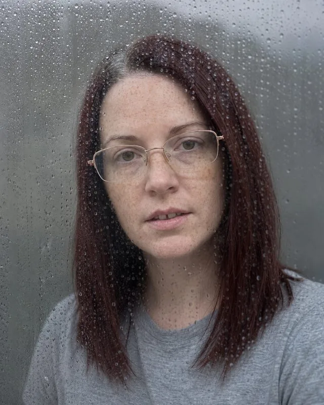 Rain window: seen through a rain-covered window with droplets sharp in the foreground, soft grey light on the face