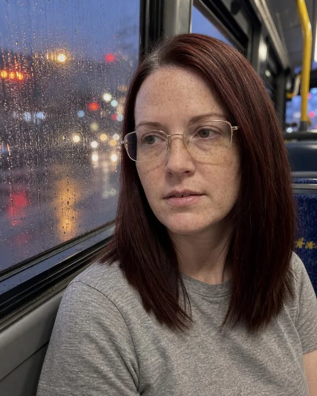Rain window: inside a bus with rain streaking the glass and city lights smeared behind, quiet expression