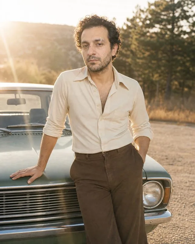 1970s: leaning on a vintage car bonnet in flared trousers and a wide-collar shirt, hazy sun flare and faded colour