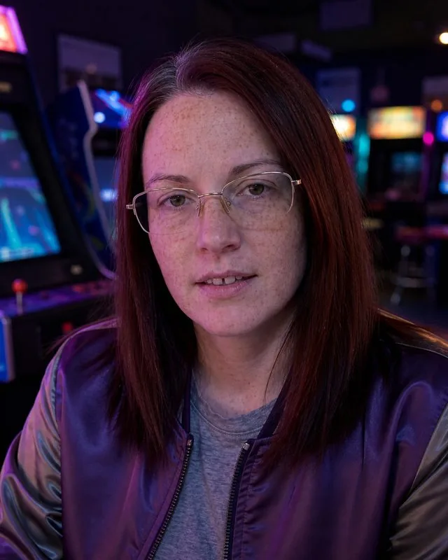1980s: in an arcade at night lit by cabinet screens, satin bomber jacket, purple and cyan glow on the face
