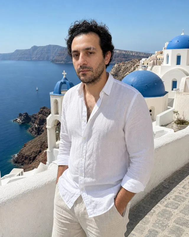 Santorini: on a whitewashed terrace above the caldera in a flowing summer outfit, blue domes behind, hard bright sun