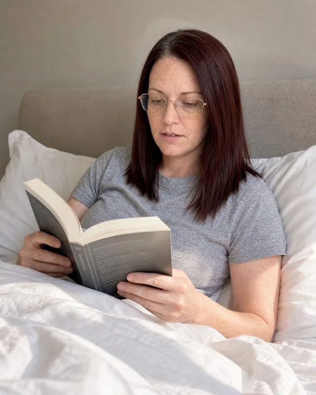 Slow weekend: reading in bed with white linen sheets and morning light across the duvet, soft neutral tones