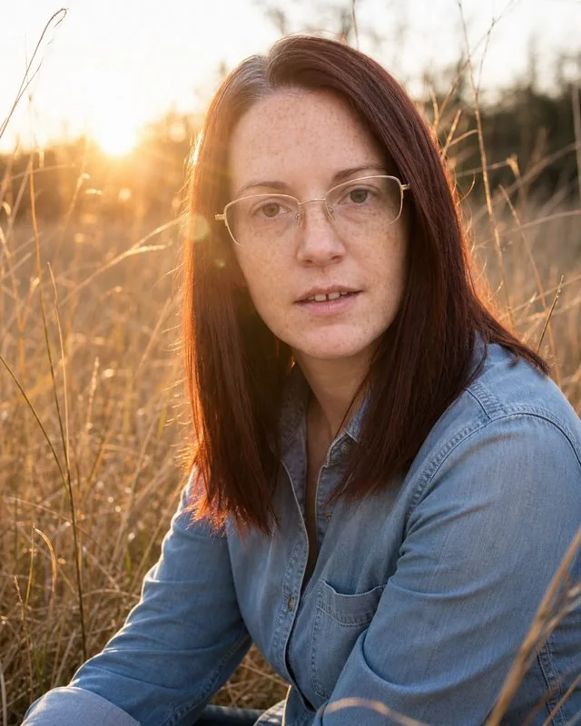 Golden hour: sitting on grass in a field of tall dry stems, faded denim shirt, low sun flaring into the lens from behind