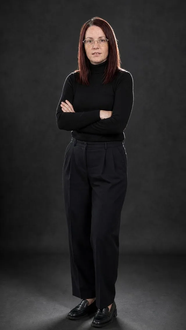 Editorial full figure: a head-to-toe frame with arms crossed on a charcoal backdrop, black turtleneck and pleated trousers