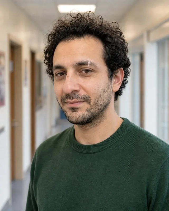 Teacher: a head-and-shoulders frame in a school corridor, plain green jumper, soft even light, welcoming smile