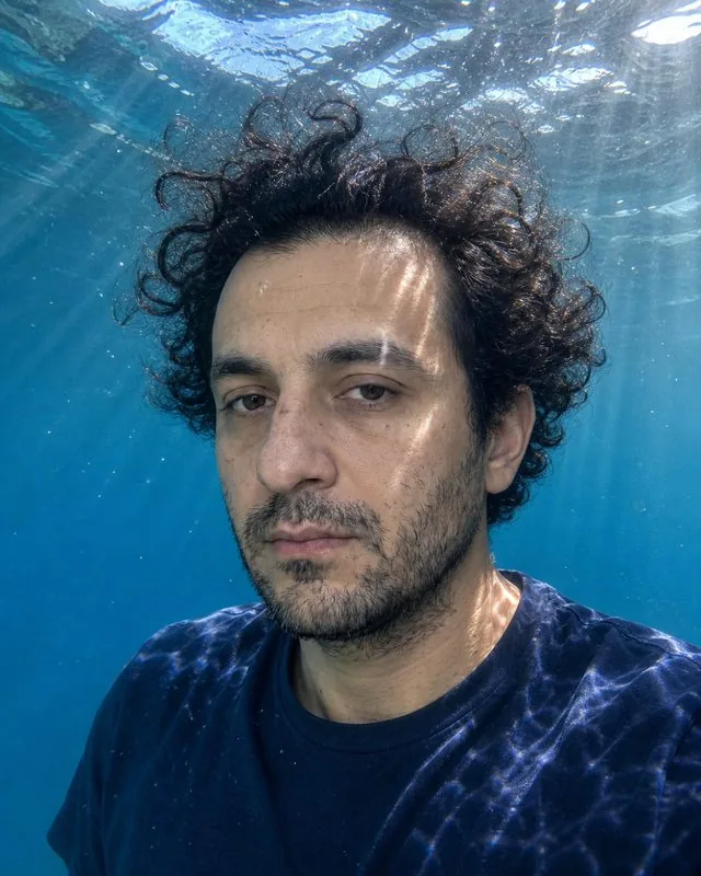 Underwater: submerged in clear blue water with hair and fabric floating, sunbeams cutting from the surface