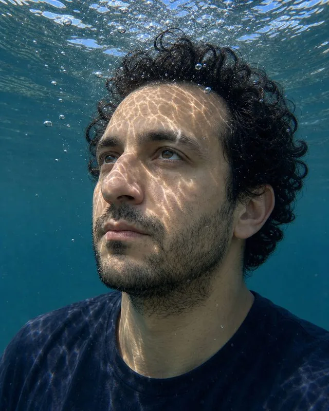 Underwater: just below the surface looking up, rippling light patterns across the face and bubbles rising