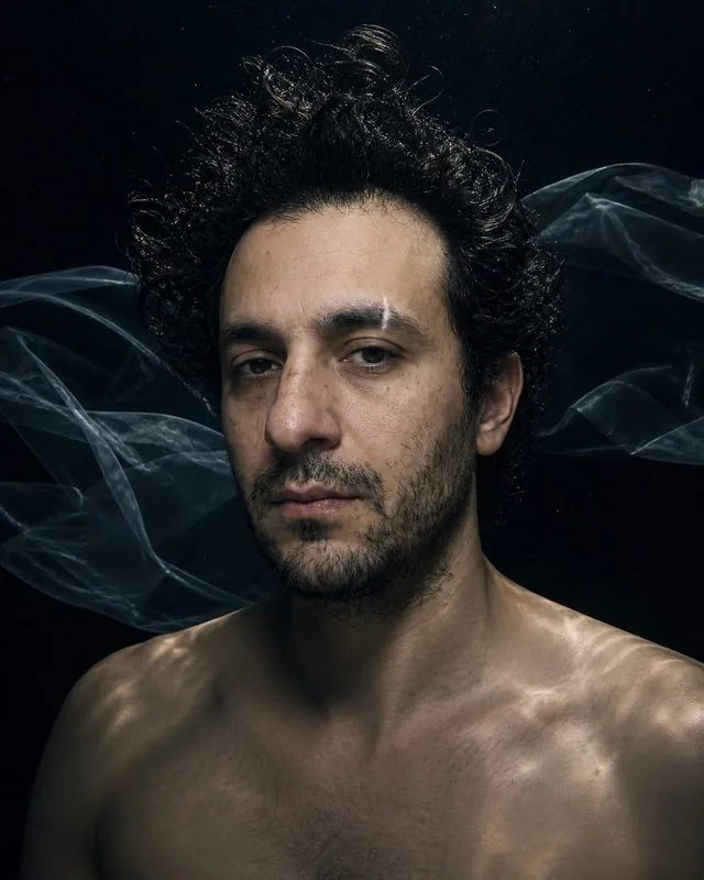Underwater: in a deep pool with dark water behind and a single hard light from above, fabric drifting