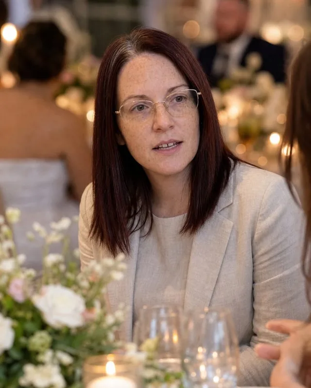 Wedding guest: seated at a decorated table with flowers and glassware, linen suit, candid mid-conversation, soft candlelight